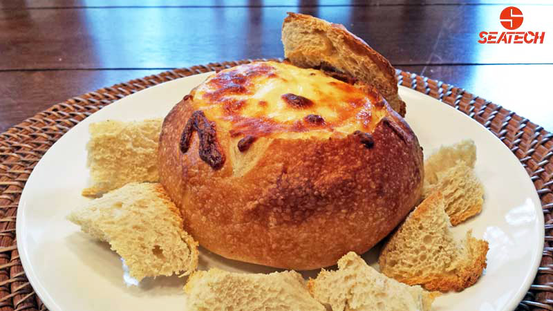 A photograph of a hollowed out loaf of sourdough bread, filled with crab dip covered with chesse and baked until golden brown.