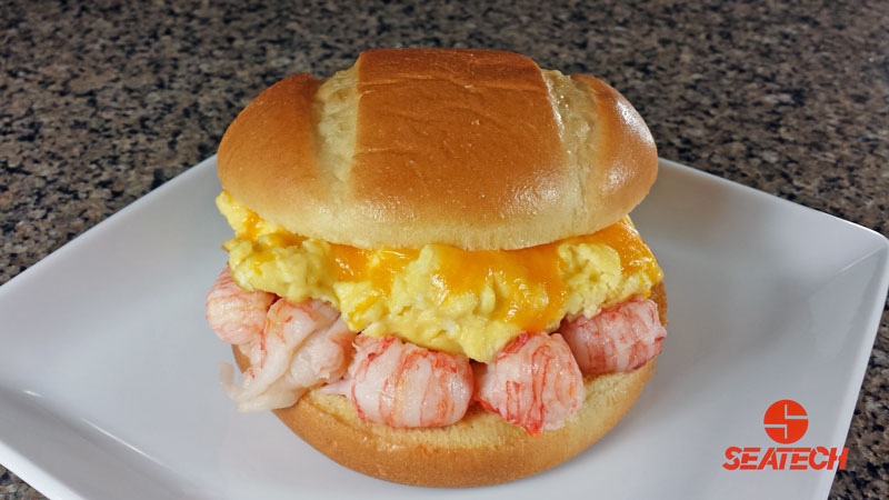 A photograph of a breakfast sandwich with langostino lobster, scrambled egss with melted cheese on a brioche bun.