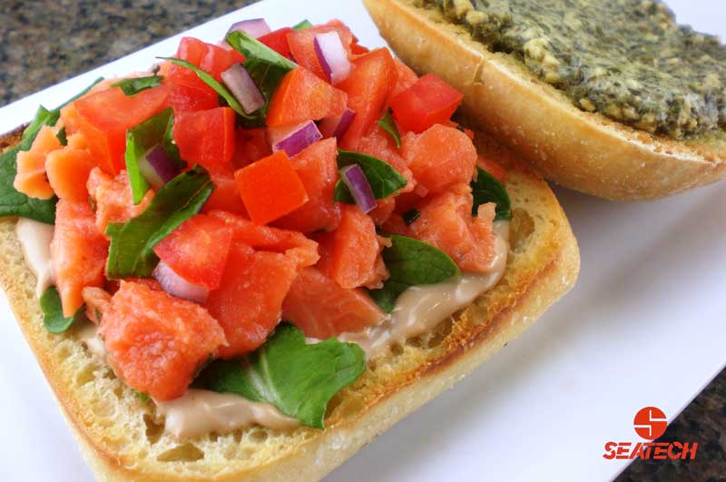 A photo of a salmon tartar bruschetta sandwich with salmon tartar, onions, luttuce, tomatoes, pesto and aoili.