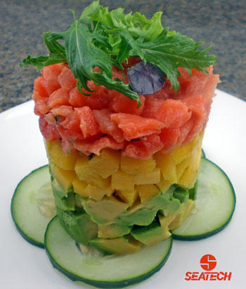 A photograph of a salmon tartar tower with a sliced cucumber base, salmon tartar layered over diced mango over diced avocado and micro greens. 