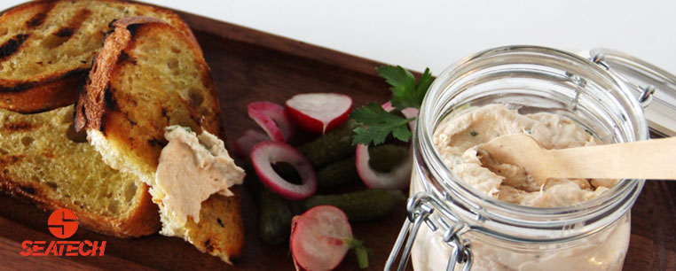 A photograph of zesty salmon dip in a jar with sliced grilled bred, red onion and cornichons